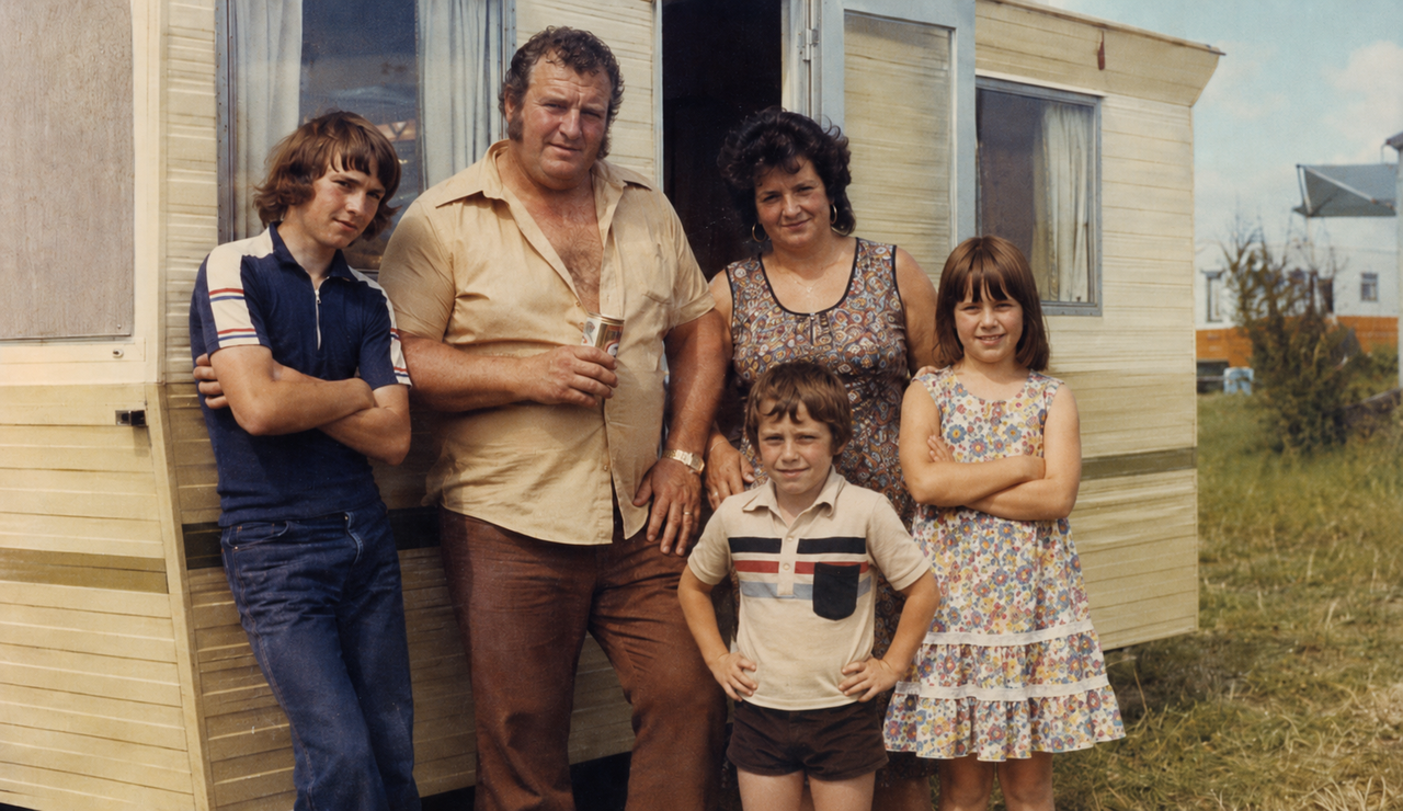Faded 1970s-style family holiday photograph outside a caravan, showing a working-class family from an earlier generation.